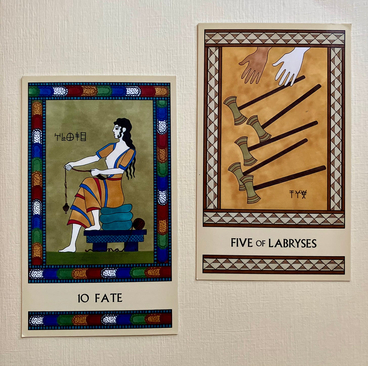 Two Minoan Tarot cards side by side on a cream background. The Fate card has a multicolor border. It pictures a Minoan woman seated on a pile of cushions, facing left, holding a drop spindle as she spins thread. The Five of Labryses is in shades of gold and tan. It shows two hands letting go of five labryses, allowing them to fall through the air.