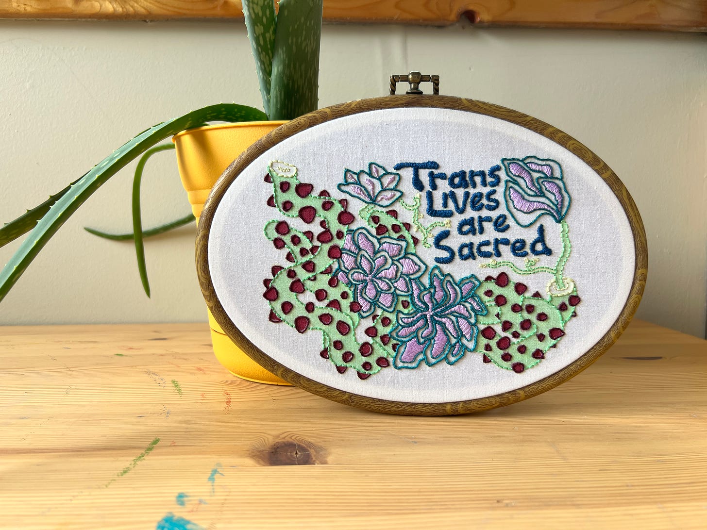 Completed mixed-media embroidery piece using the design created by Ris Wong of winding thorny vines and blossoming many-petalled flowers. The piece is leaning up against a yellow plant pot with an Aloe plant growing in it. 