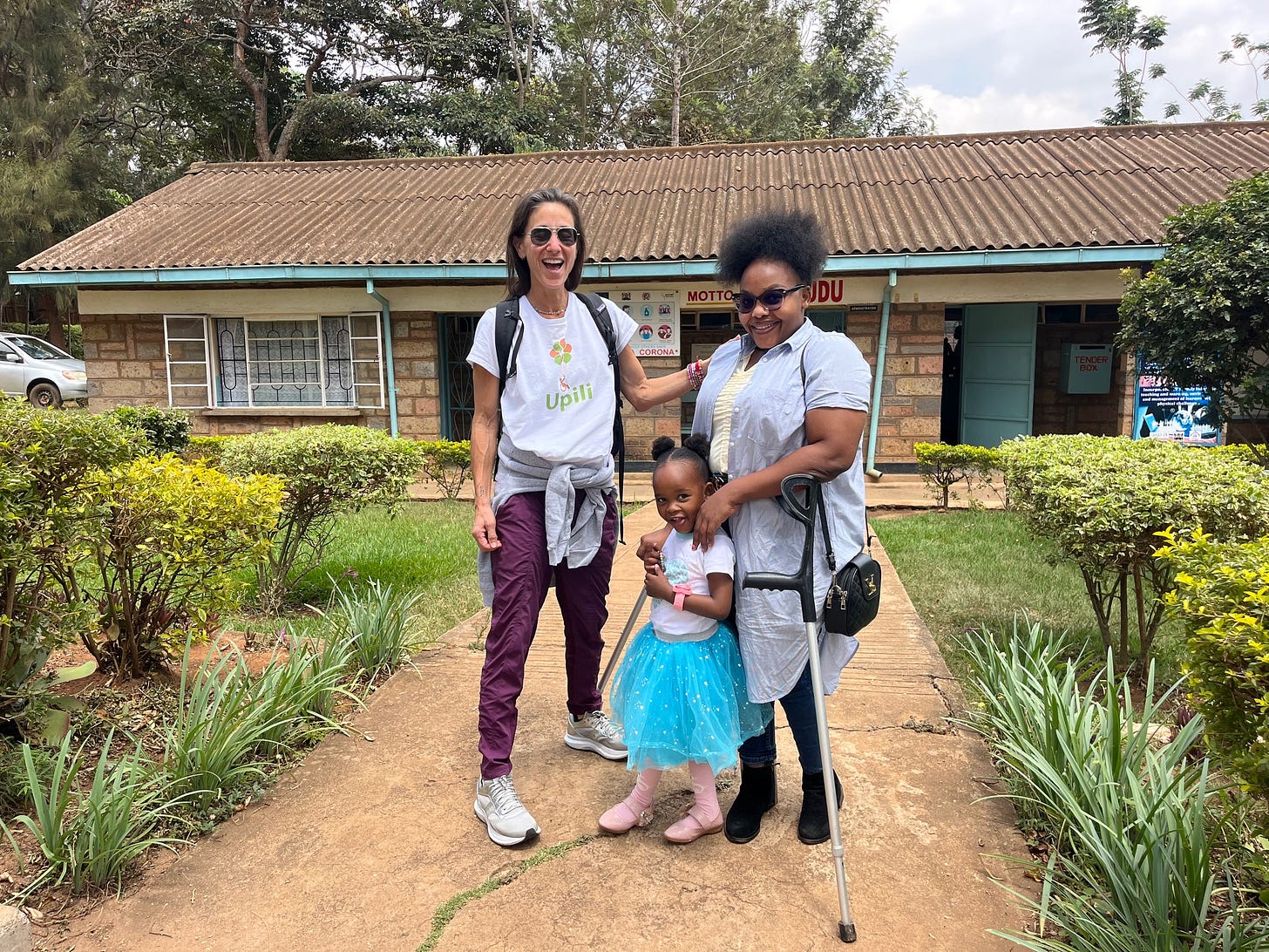 Carla stands in front of the school with a woman and her young daughter. The school is set on a grassy path