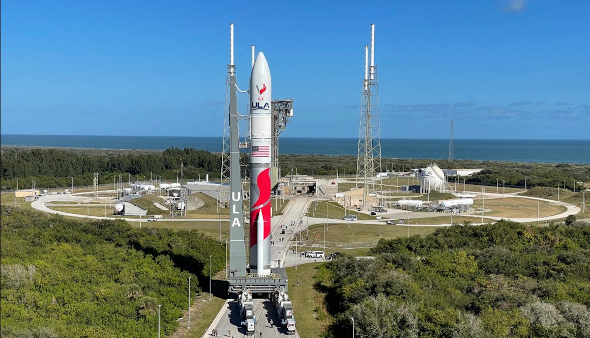 A large rocket is positioned vertically on a launch pad, surrounded by green foliage and set against a clear blue sky. Structures and equipment are visible in the background.