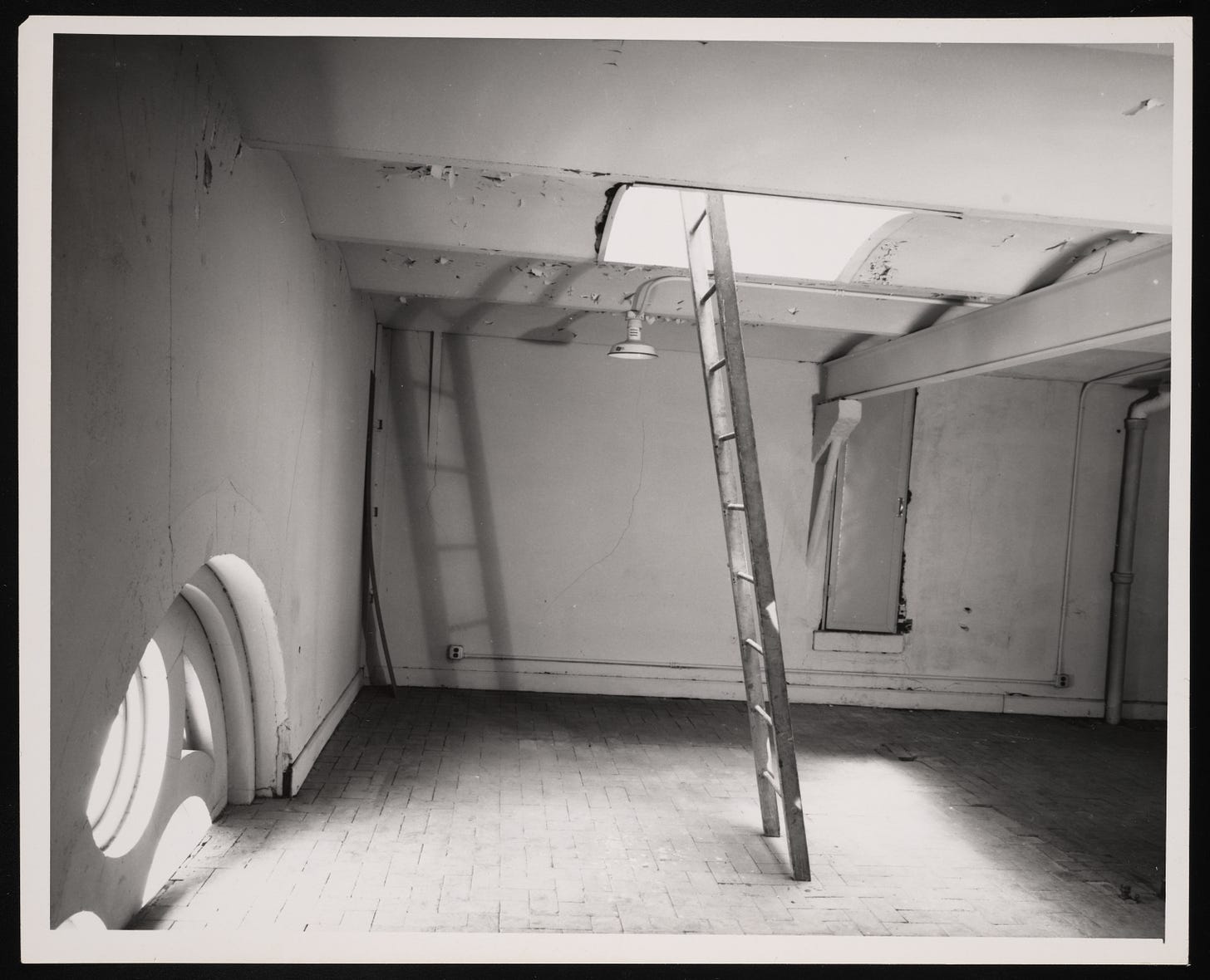 A black and white photograph of a low-ceilinged room where there are two sources of light. One is in the bottom left of the photo, where daylight streams through the top half of a trefoil window. The second is a hatch in the ceiling. Reaching up through the hatch is a wooden ladder, the top of which seems to rest against nothing.