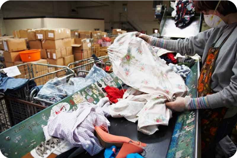 A women in a clothing sorting facility holds up a floral garment while the conveyorbelt of other clothes goes past underneath.