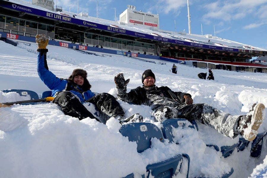 Two people wave while sitting in deep snow amid stadium seating.