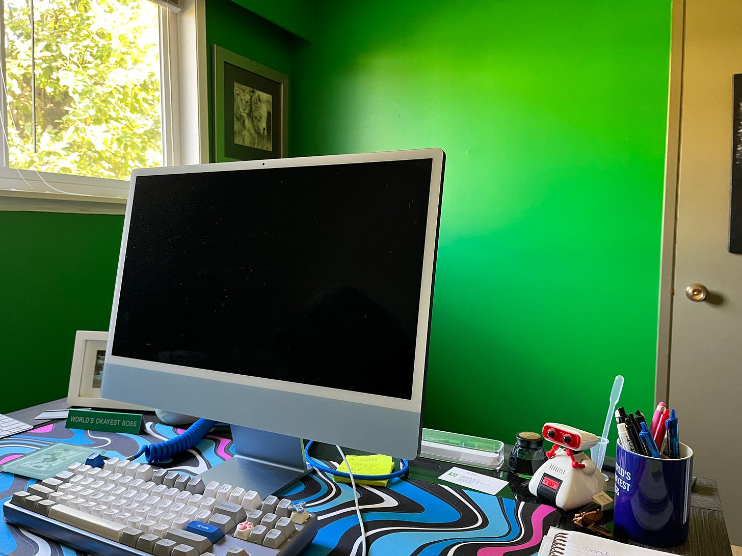 A Mac computer sits on a clean desk that also contains a tiny robot, a fountain pen, and small papers