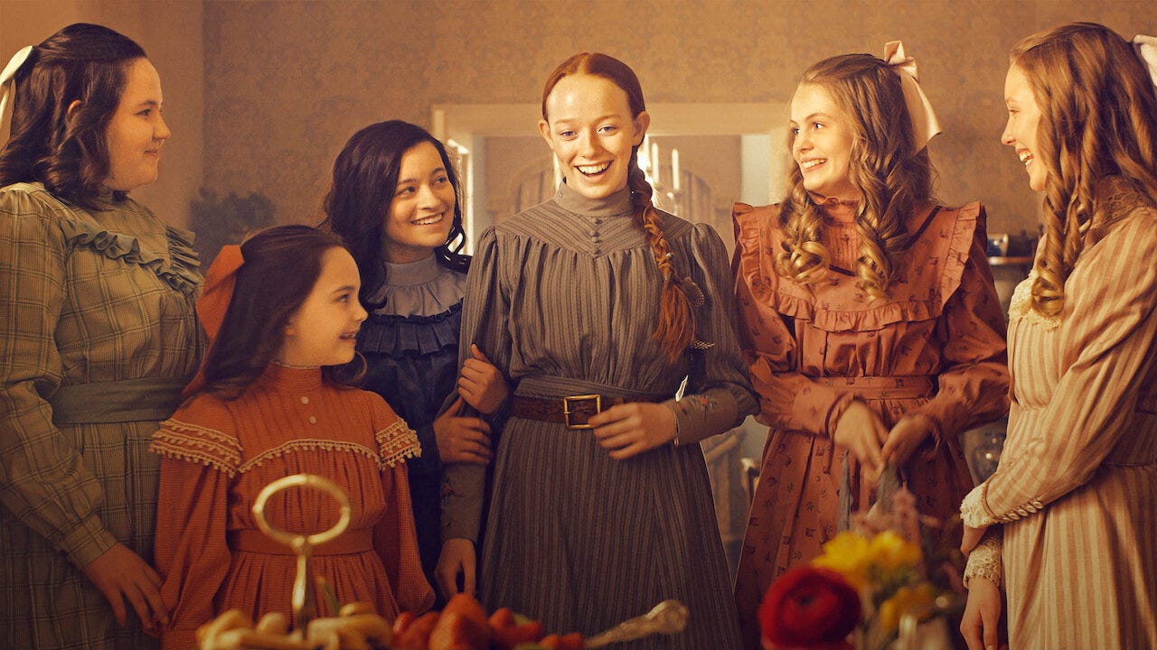 Girls in 1890s outfits standing together, smiling around a table (offscreen) of food