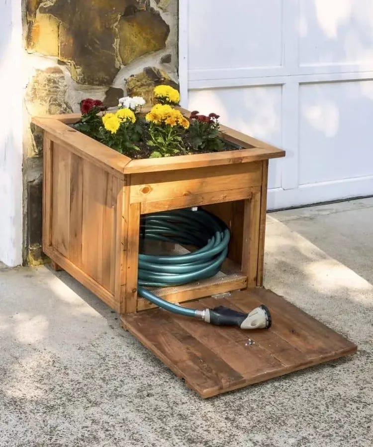 A photo of a large square wooden box planter with yellow, white and red flowers planted in the top and the space underneath the flowers in the base of the box used for storing the hosepipe.