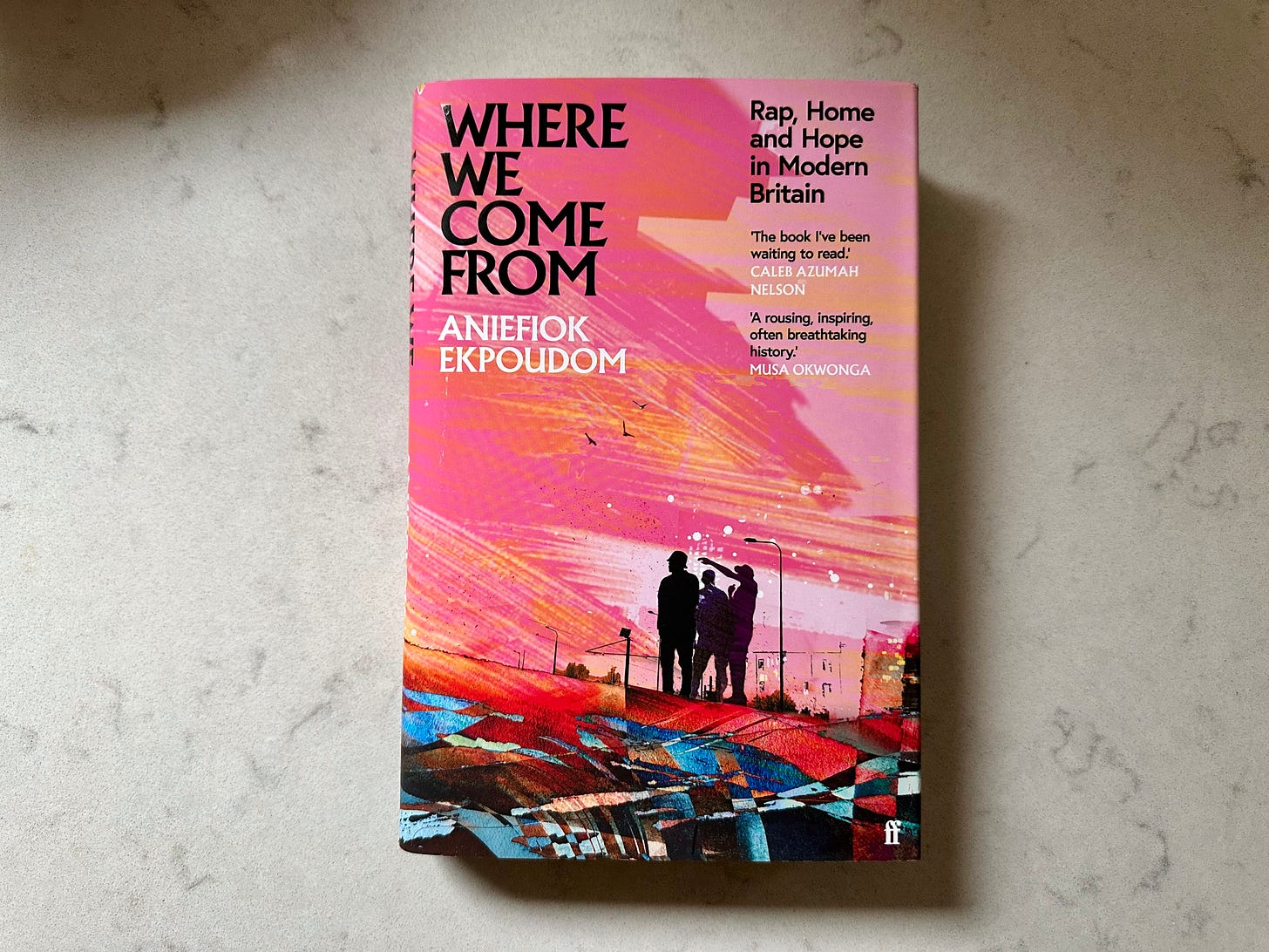 Picture of a book titled "Where We Come From" by Aniefiok Ekpoudom, on a white marble table. The cover has a contemporary pink cover with broad brush strokes showing a small group of rappers in silhouette.