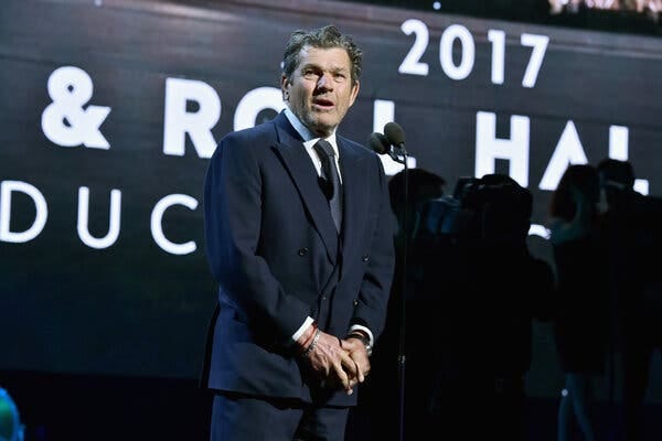 Jann Wenner in a navy suit standing in front of a microphone.