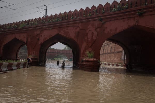 Two people wade through hip-deep water under a bridge.