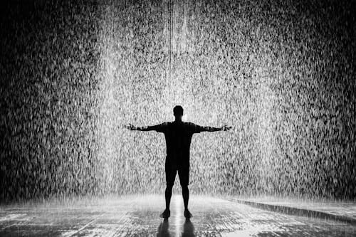 Free A person joyfully standing in the rain with arms wide open in a dramatic black and white scene. Stock Photo