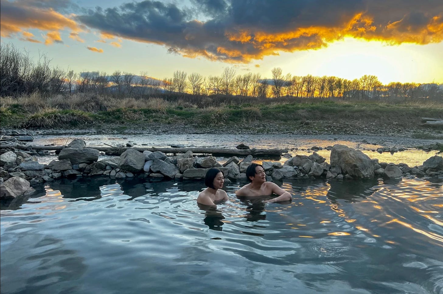 A Japanese man and woman are immersed in an outdoor hot spring with a beautiful sunset in the background.