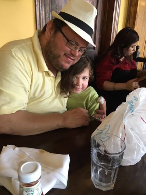 A bearded, bespectacled man, wearing a yellow shirt and an off-white hat with a dark band, smiles tenderly as he sits and holds on his lap a sweetly-smiling little girl wearing a green top. They are in a restaurant.