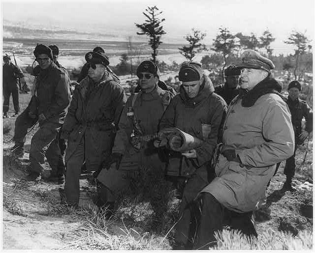 Black and white photo of (from right to left) General Douglas MacAurthur, Major General Courtney Whitney, Lieutenant General Matthew B. Ridgway and Major General William Kean in cold weather clothing on a snowy hillside near Suwon, Korea in January 1951. There are several other soldiers in the background as the generals, standing shoulder to shoulder, look together across the battlefield. Black and white photo of (from right to left) General Douglas MacAurthur, Major General Courtney Whitney, Lieutenant General Matthew B. Ridgway and Major General William Kean in cold weather clothing on a snowy hillside near Suwon, Korea in January 1951. There are several other soldiers in the background as the generals, standing shoulder to shoulder, look together across the battlefield.