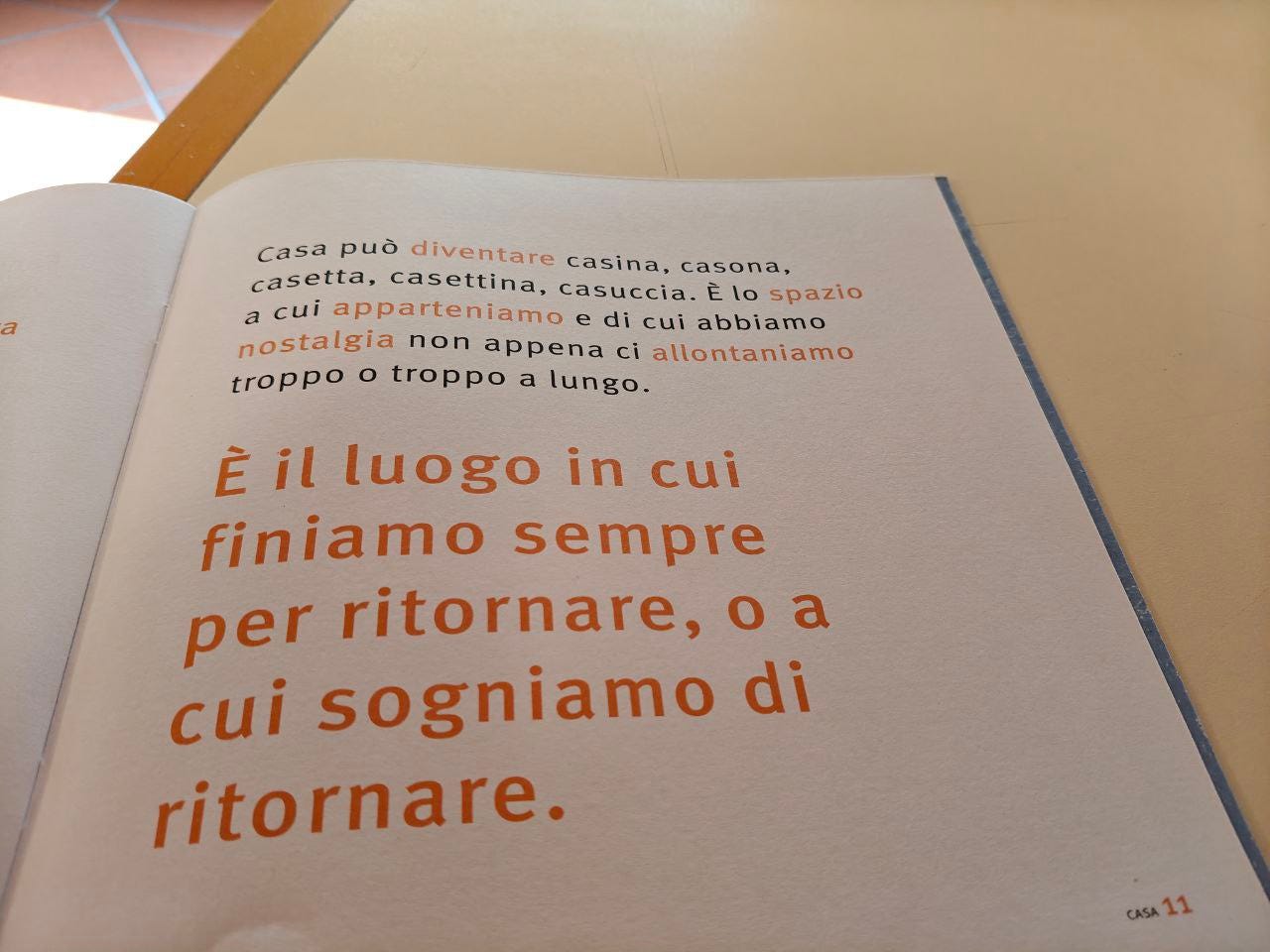 Una pagina della parola Casa delle Scatoline di effequ con questa scritta in arancio e nero: "Casa può diventare casina, casona, casetta, casettina, casuccia. È lo spazio a cui apparteniamo e di cui abbiamo nostalgia no appena ci allontaniamo troppo o troppo a lungo. È il luogo in cui finiamo sempre per ritornare, o a cui sogniamo di ritornare. La pagina è appoggiata su un tavolo di legno bianco sporco e nell'angolino in alto a sinistra si intravede un pezzetto di pavimento in cotto.