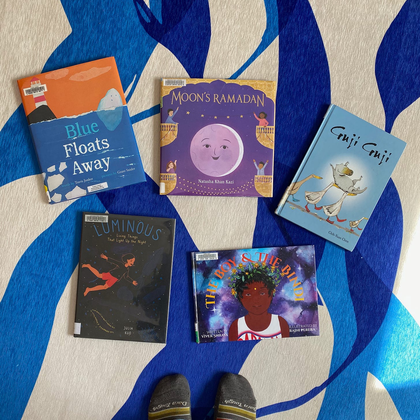 Five picture books arranged in a semi-circle on a blue and white rug: Blue Floats Away, Moon’s Ramadan, Guji Guji, Luminous, and The Boy and the Bindi.