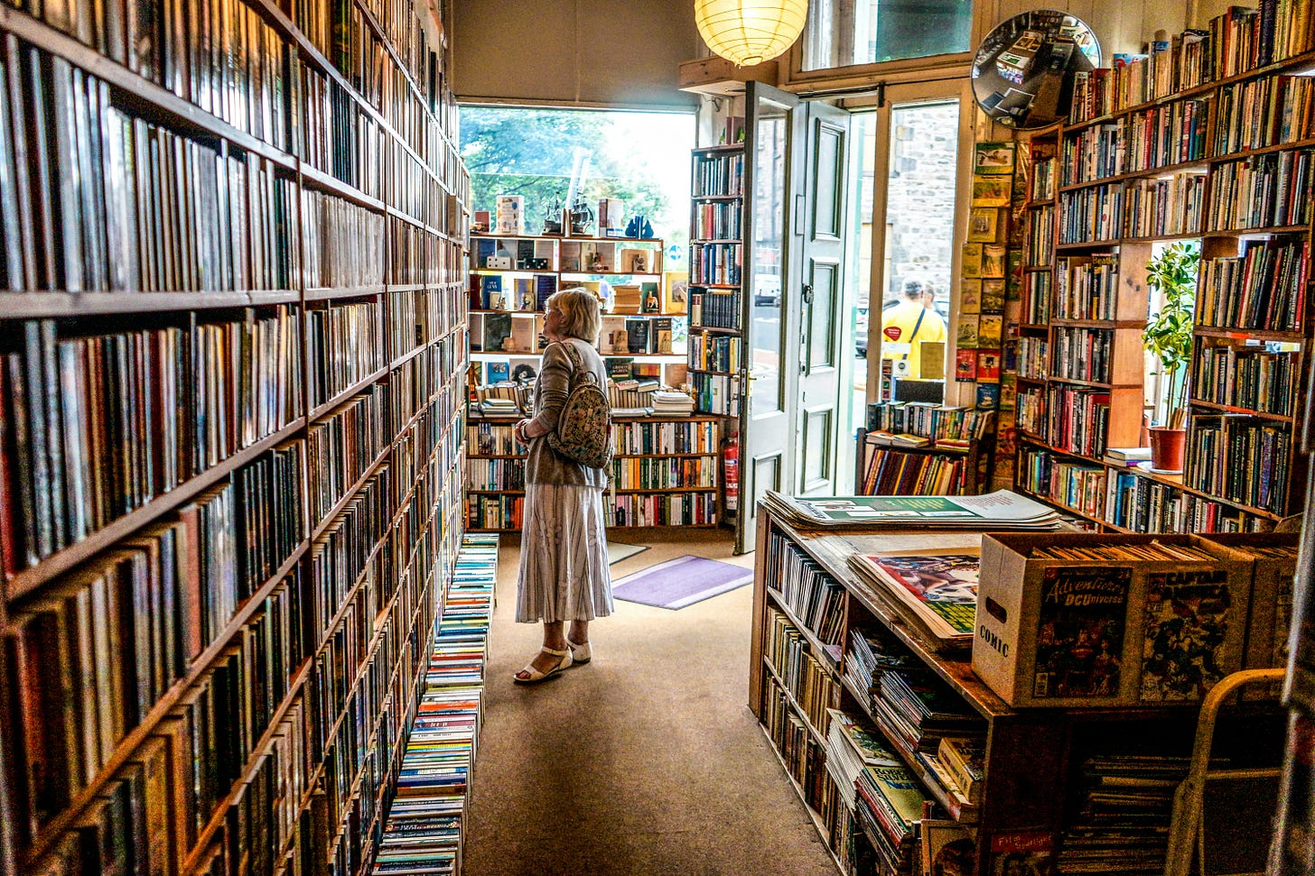 a book store filled to the brim with shelves of books. the books look worn and used. a woman with grey hair, a blue jacket, and a grey dress stands in the center of the photo, looking left toward the shelves. a lower table of books stands to the right, behind her.