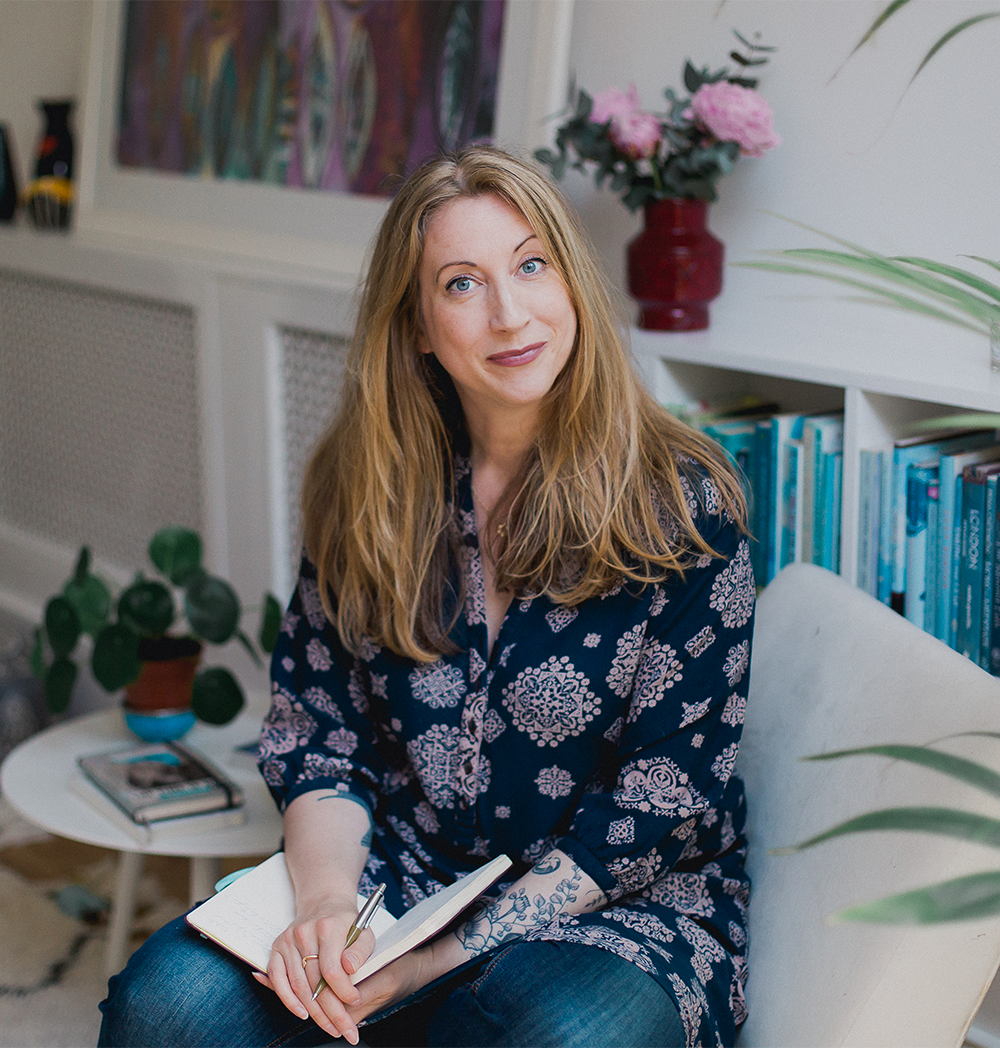 Susannah is a white woman with long, dark blonde hair, sitting on a sofa in front of a colourful bookcase and holding a notebook and pen in her lap. 