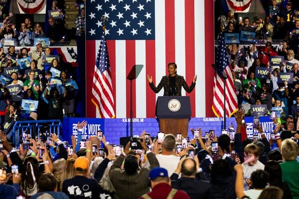 Michelle Obama, the former first lady, speaks at a rally for Vice President Kamala Harris. A large American flag is behind her and she is surrounded by supporters holding “Harris Walz” signs.