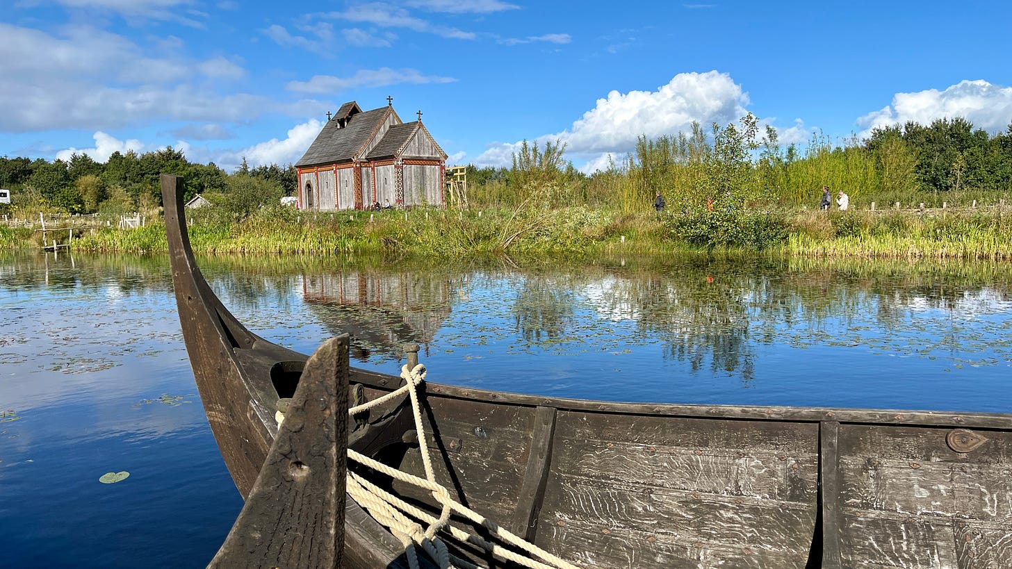 wooden viking rowboat in foreground, on water, with a medieval Viking church in the background in a field