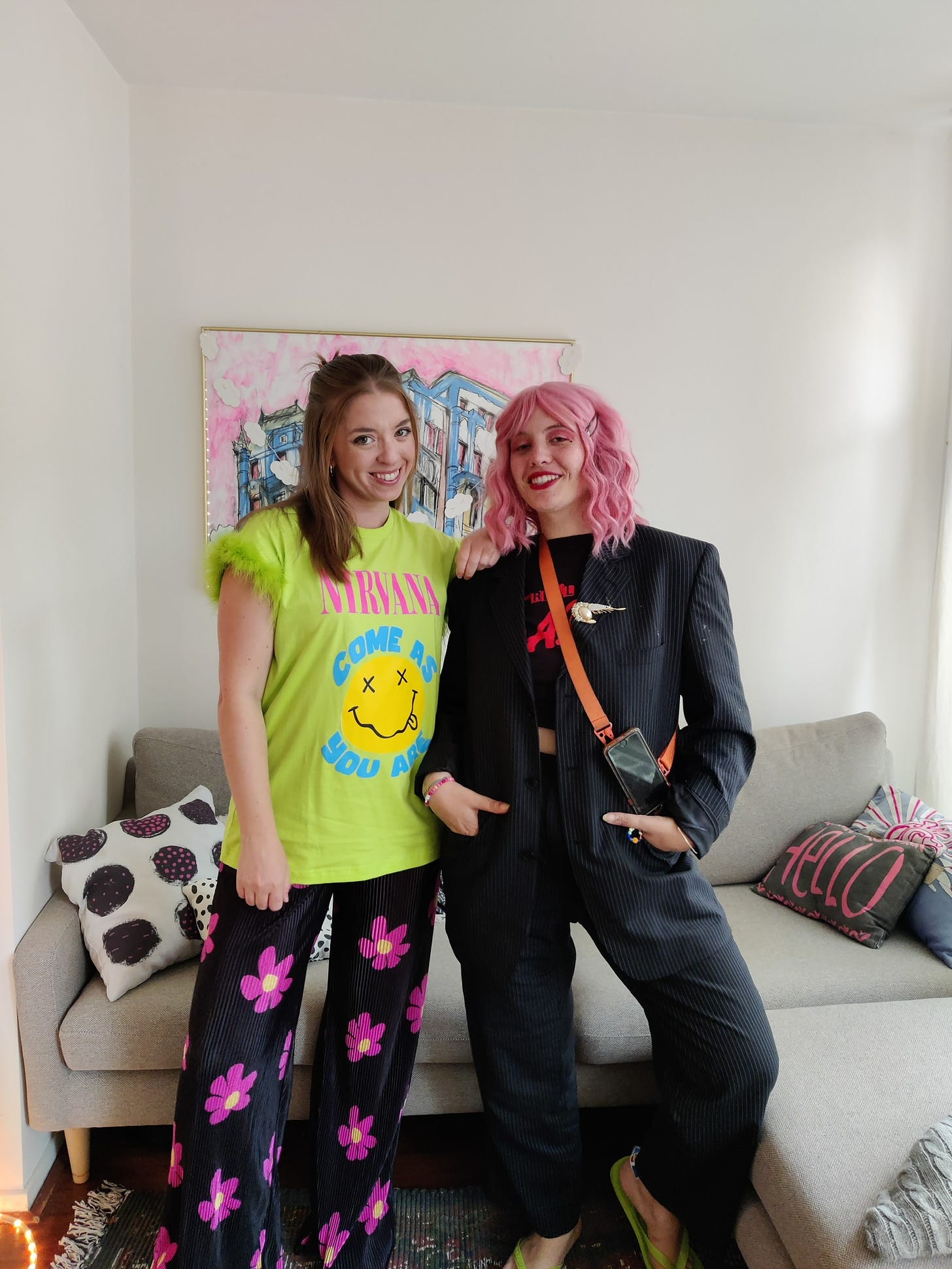 Kelly and Larissa smiling, smizing, thriving. Kelly wears a neon green tee and pink and black floral pants. Larissa wears a black ensemble and pink wig.