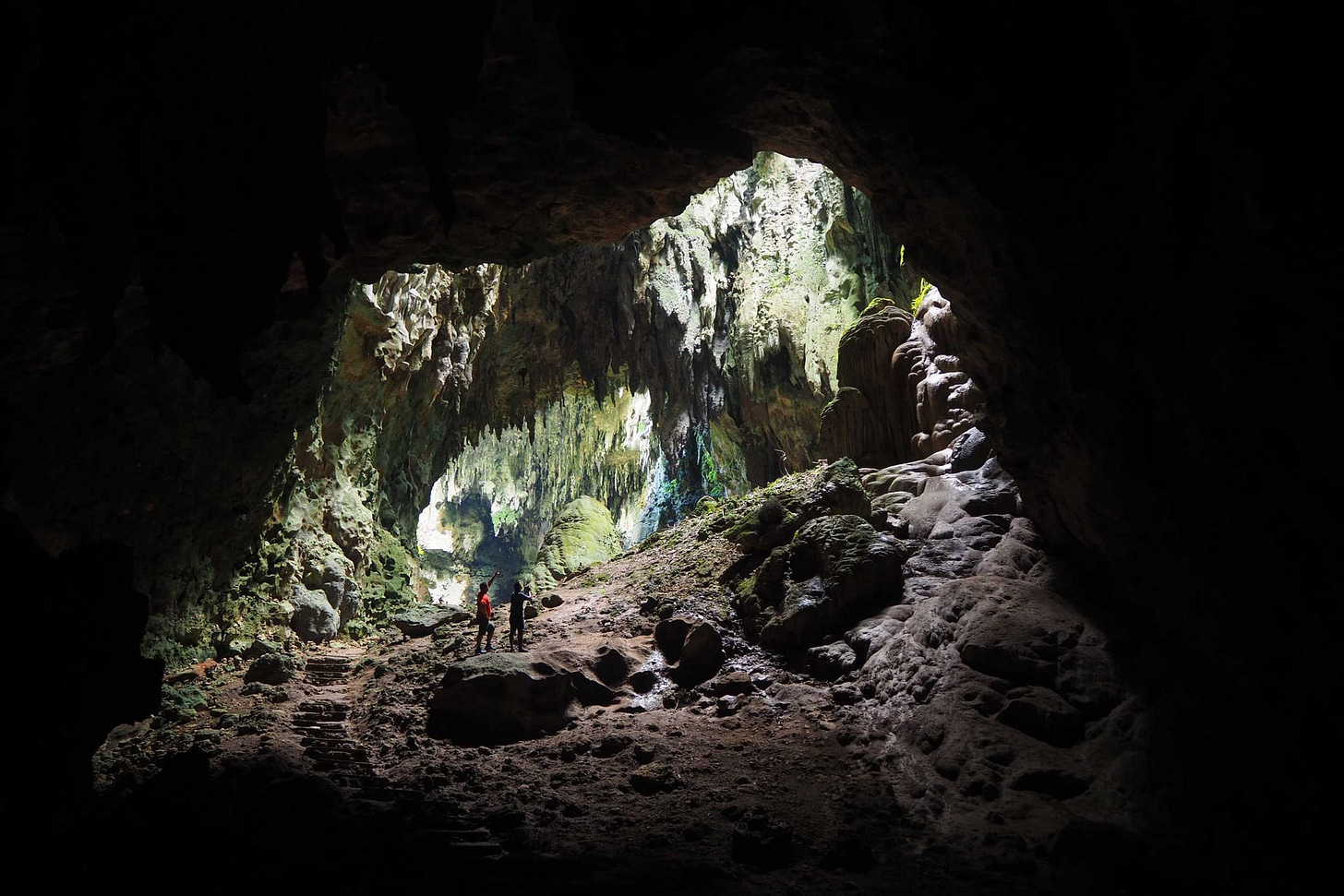image of two people within a very large cave with green color on stalactites in the background