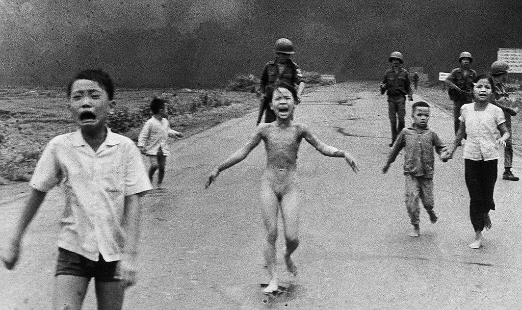 Group of children and soldiers moving on foot away from a distant cloud of smoke rising from the ground. Several children are crying and one in the center is also naked as she runs toward the camera.