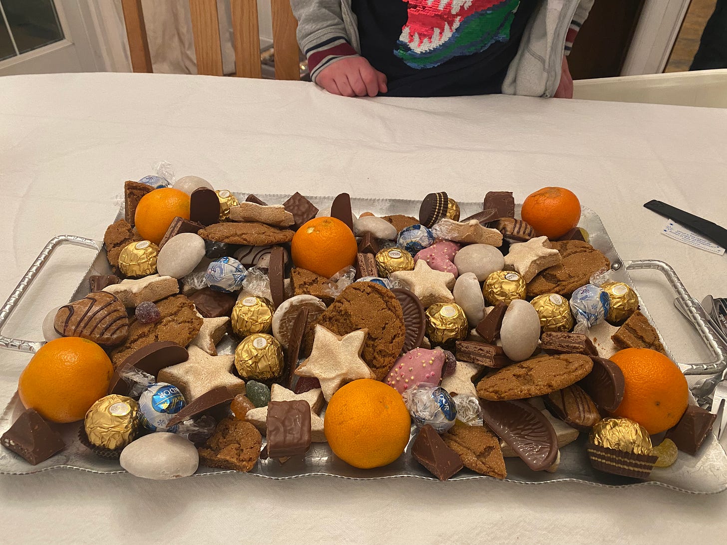 A large platter of chocolates, German cookies, and mandarin oranges. In the background a child wearing a sparkly dinosaur shirt rests his hand on the table.