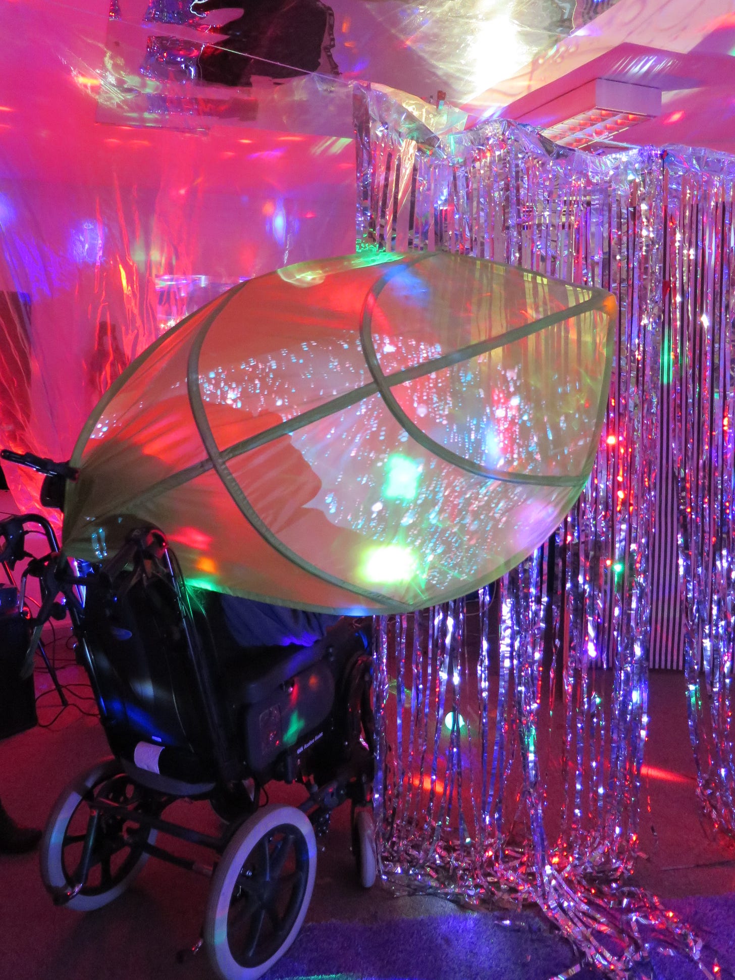 A day center attendee sits in a motorized wheelchair, back to the camera, surrounded by a a room infused with neon pink light, glittery plastic silver fringe, and big swaths of fabric suspended from the ceiling in a multisensory immersive theater.