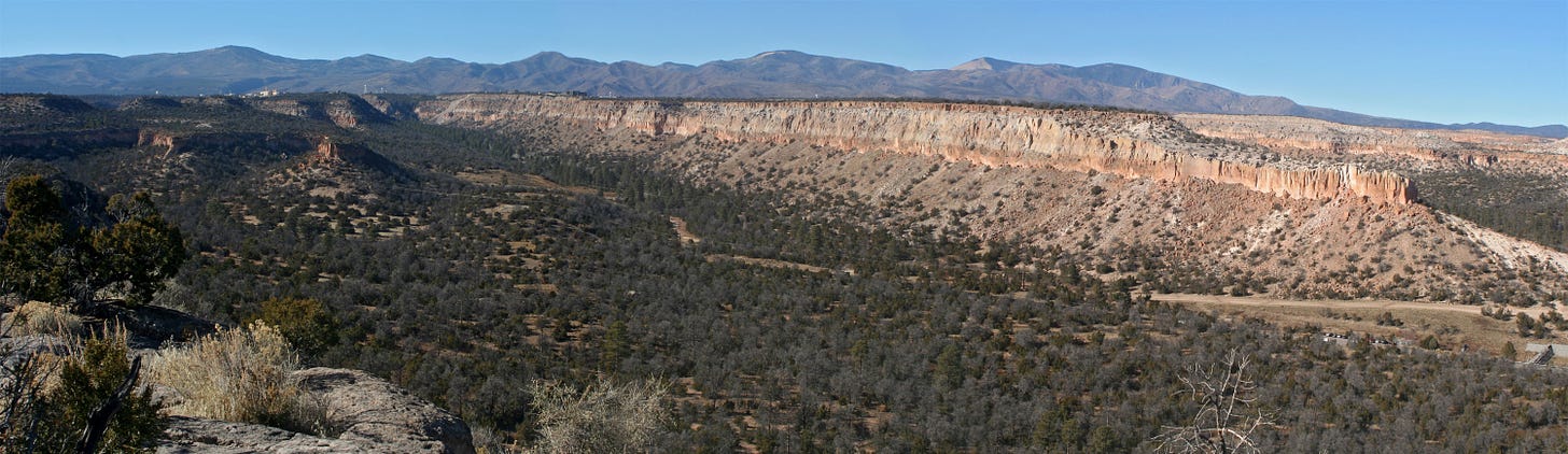 A wide photograph of mesas in Northern New Mexico, with Los Alamos visible faintly in the far left of the picture.