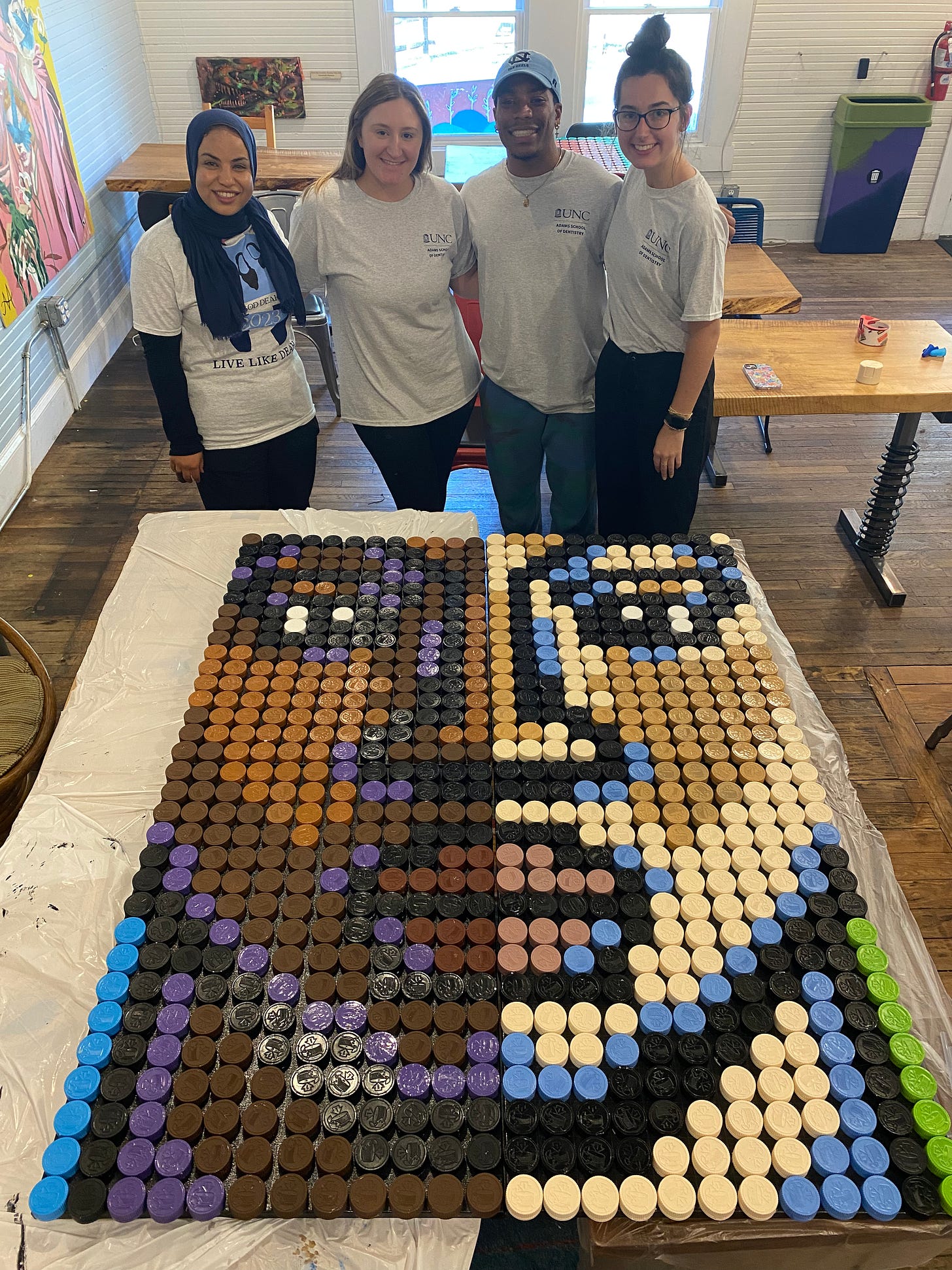 3 UNC students and 1 community member standing behind a two-tone face we created with spray painted pill bottle caps.