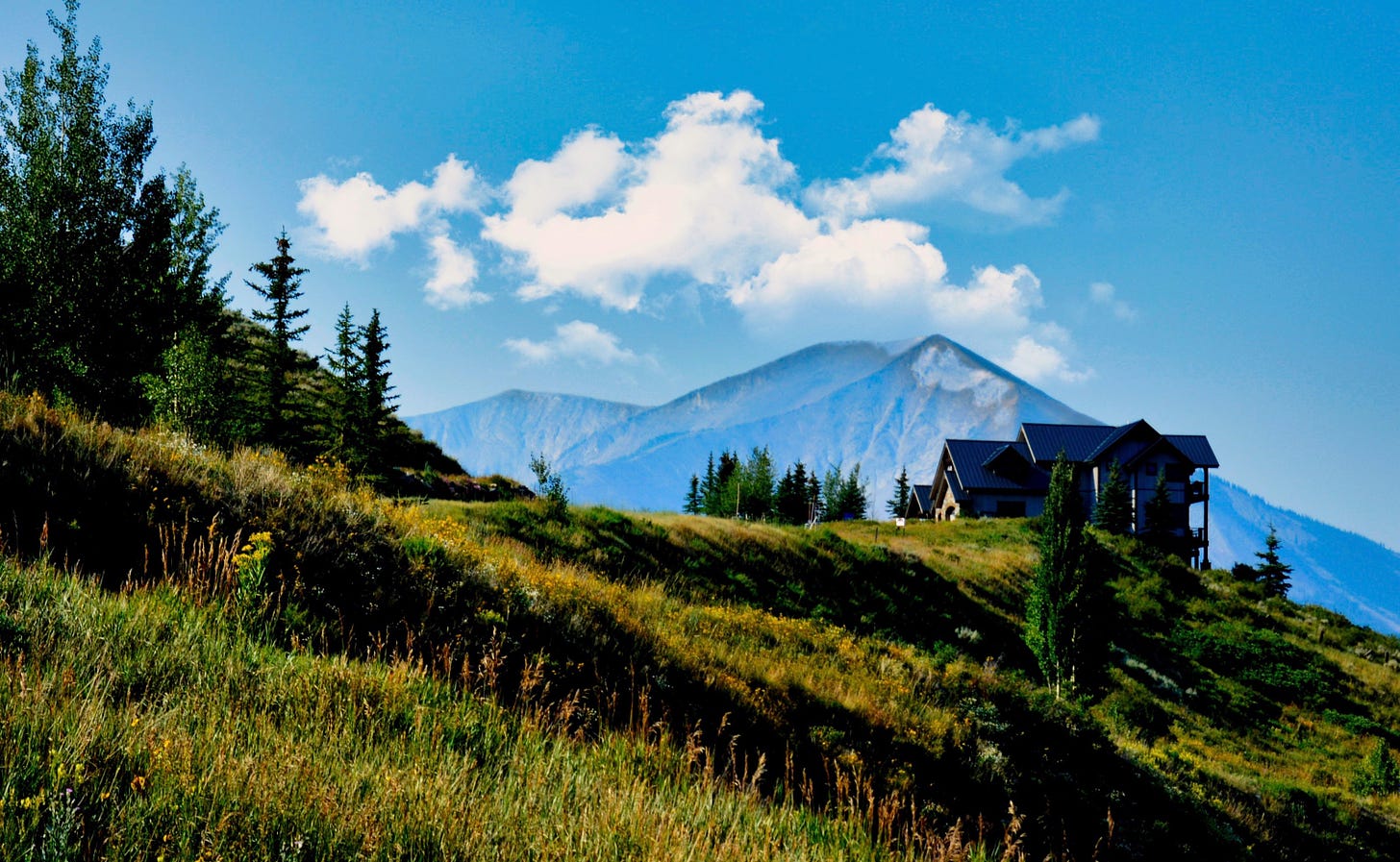 A small cabin on a hillside of the Colorado Rocky Mountains with a mountain in the background, blue skies, and white puffy clouds