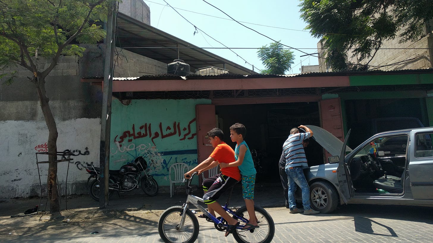 Two young boys ride a bicycle past a man repairing his car