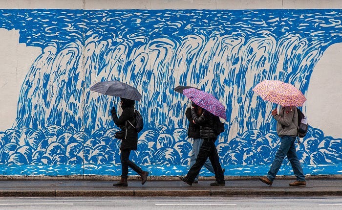 People walking by a colourful wall carrying different umbrellas