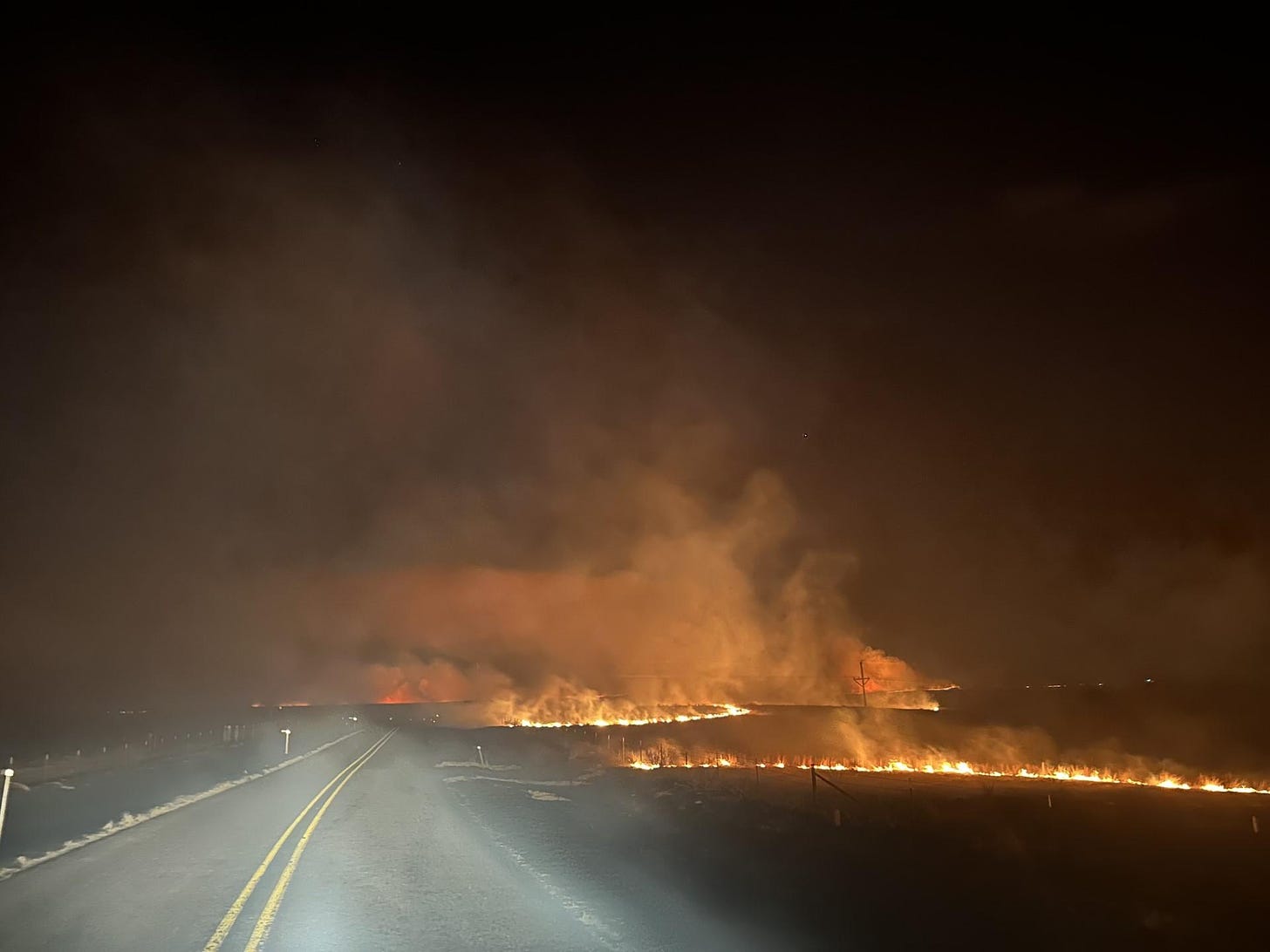 Photo depicts medium sized flames and smoke crossing a two lane road at night.