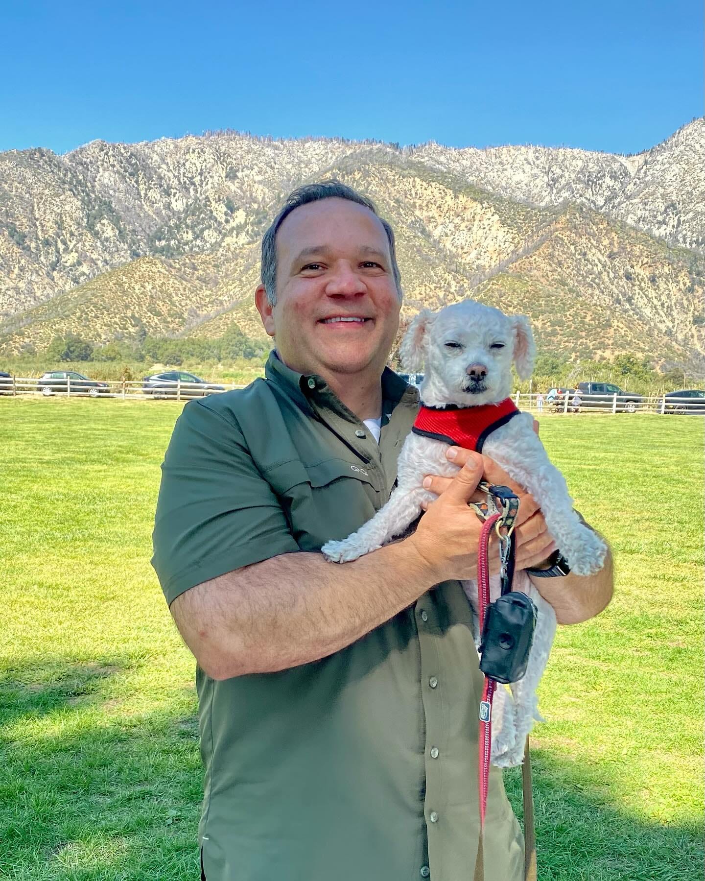 Mickey is wearing a green shirt, and holding Ellie. They are standing in a green field. There's a mountain and bright blue sky behind.