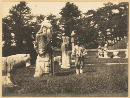 A Black and White photo of a Joseon era tomb with the tombkeeper standing next to one of the stone guardians