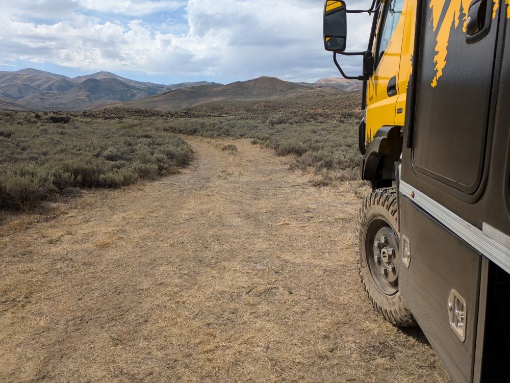 Looking past the front corner of the big yellow truck to the dirt road in the desert and the mountains in the background