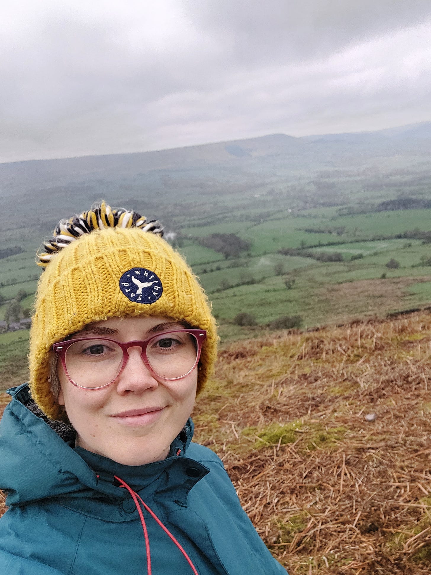 A selfie of Janelle, smiling at the camera wearing a yellow hat and pink glasses. She is white with brown eyes. Behind her is a misty view of fields and a distant fell