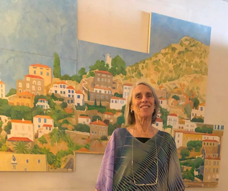 A woman stands smiling with glasses hanging around her neck and behind her is a large wall installation of a multi-panel oil painting with houses on a hillside and blue sky.