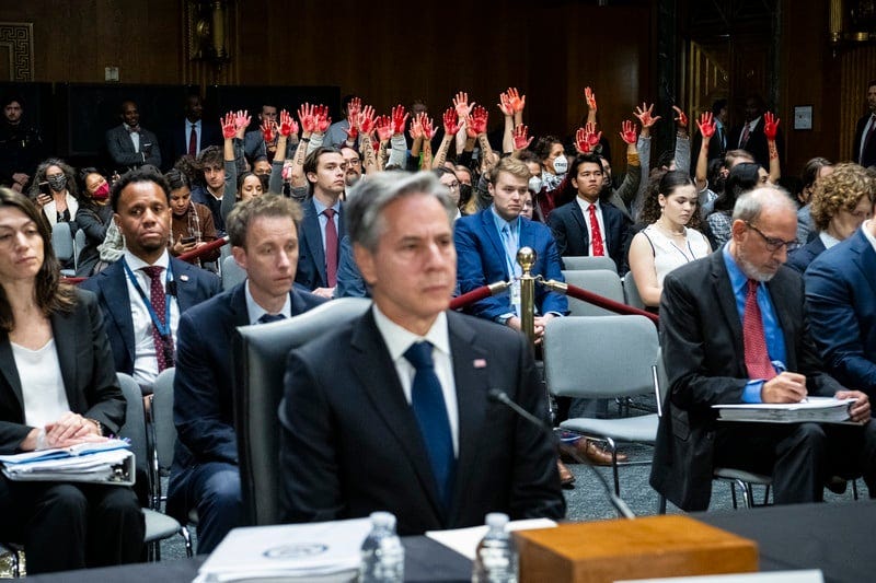 Man in a suit is seated while audience behind him has their hands raised and painted red Man in a suit is seated while audience behind him has their hands raised and painted red