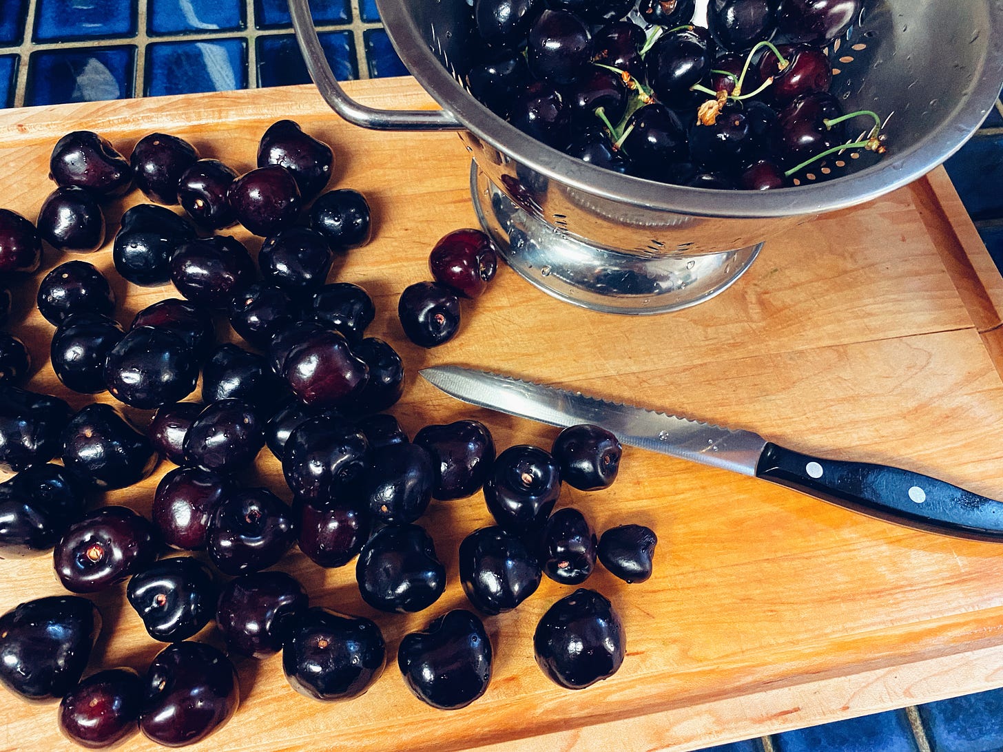 a photograph of santina cherries on a wooden cutting board. There is also a silver colander full of cherries, and there is a knife on the cutting board.