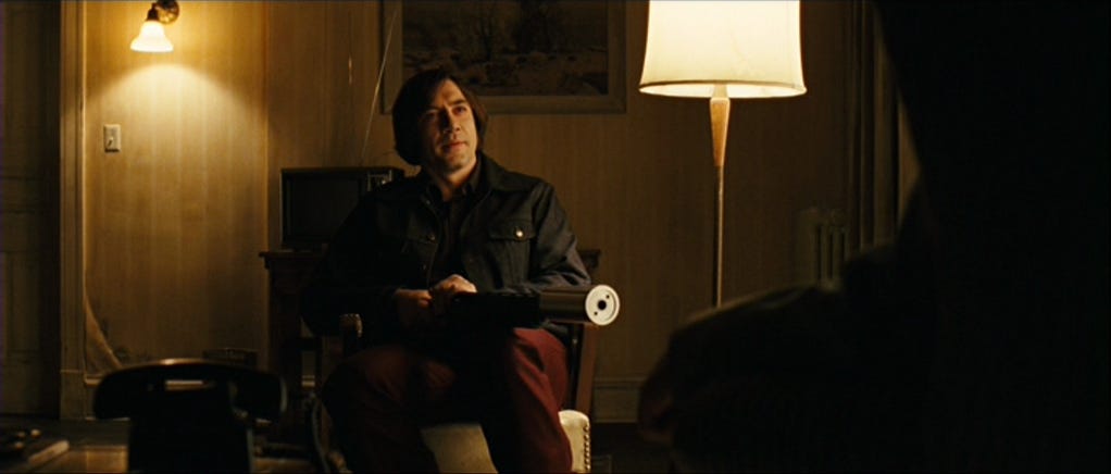 Anton Chigurh holds a shotgun with a large, cylindrical silencer across his lap.