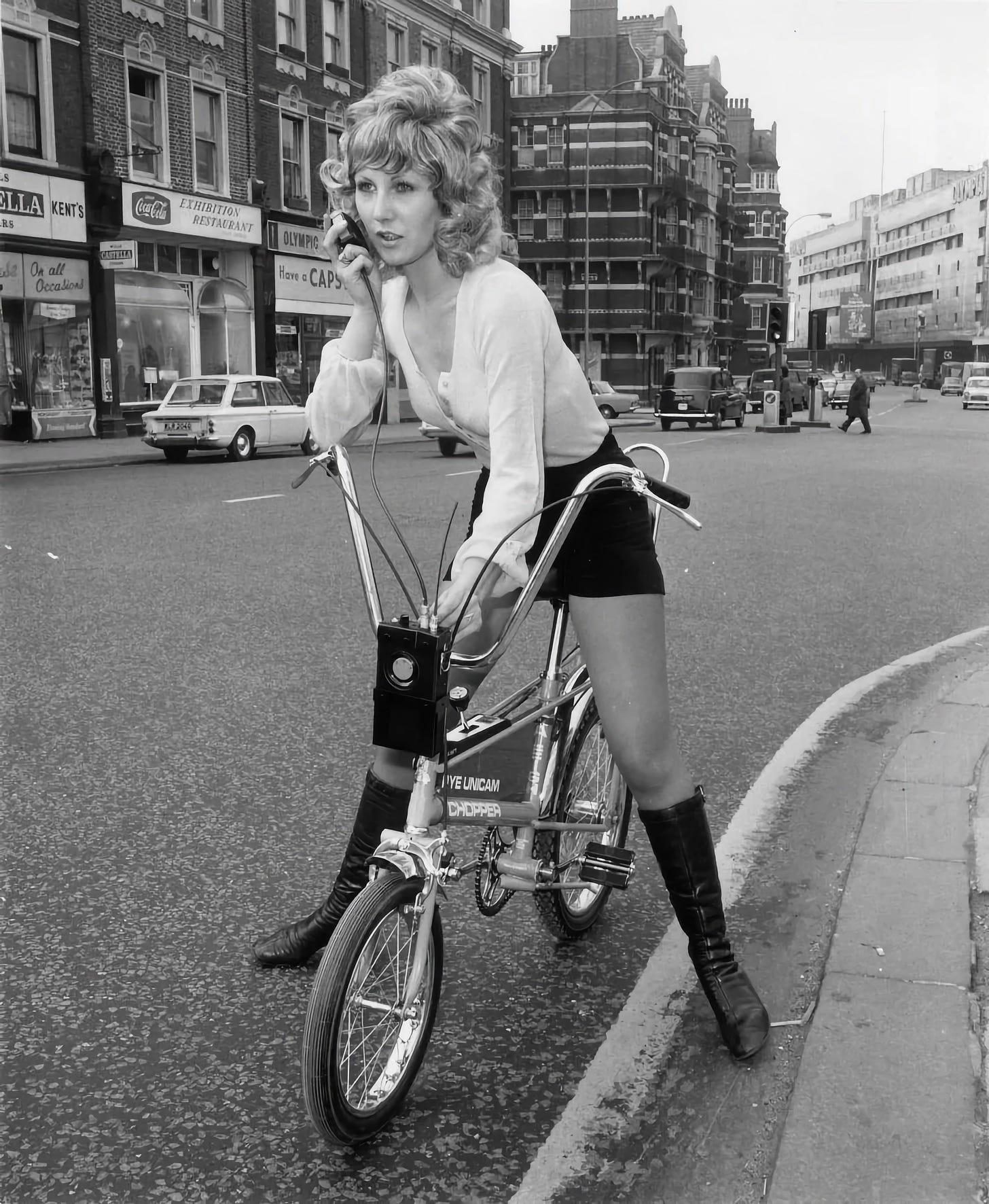 A black and white photo pop a lady somewhere in London sitting astride a Raleigh chopper while using a radio telephone.