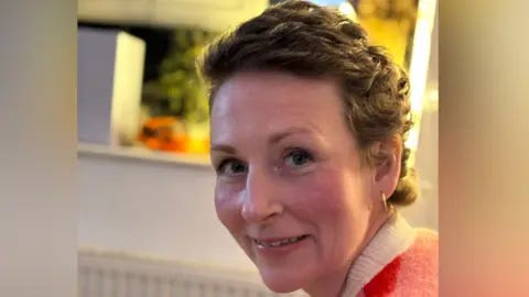 Claire O'Shea Claire is in a home and is looking over her left shoulder at the camera while smiling slightly. She has short brown wavy hair and is wearing gold hoop earrings. She has a coral coloured jumper with with a white collar and behind her is an out-of-focus kitchen.