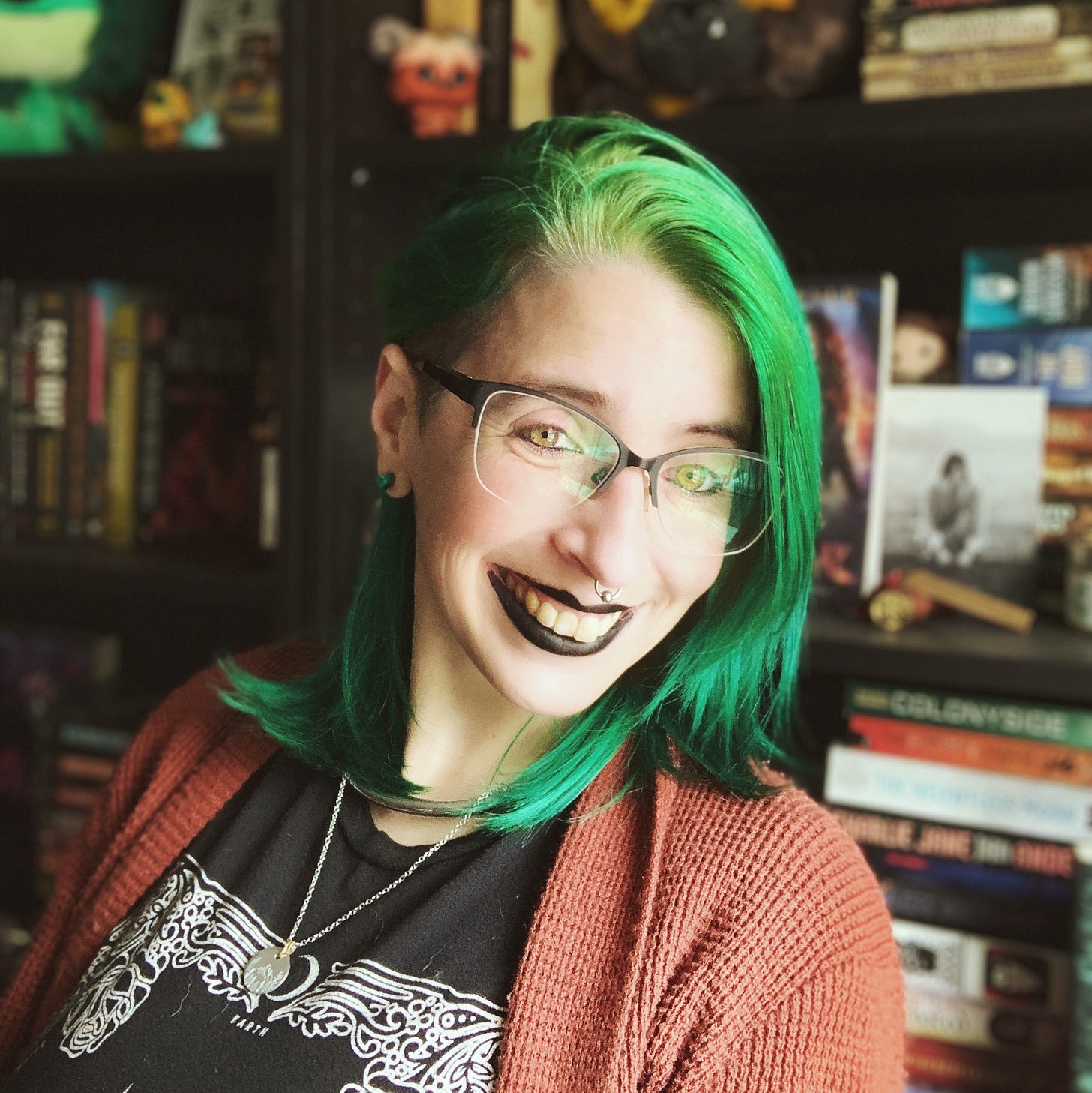 Jen stands in front of her bookshelves smiling. She is wearing black lipstick, her green hair is swepts over, and she's wearing an orange cardigan. 