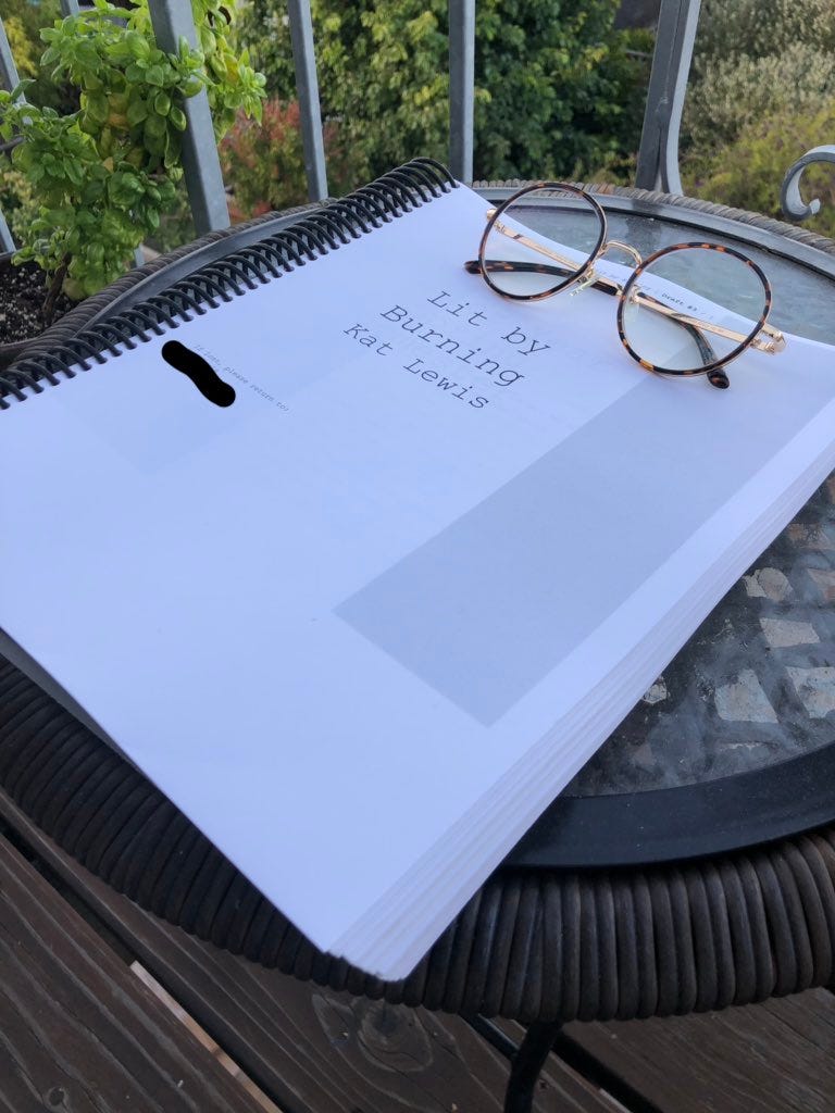 A photo of the 340-page printed manuscript of Kat's novel, Lit by Burning. It sits on a glass balcony table with her round, tortoise shell glasses atop it.