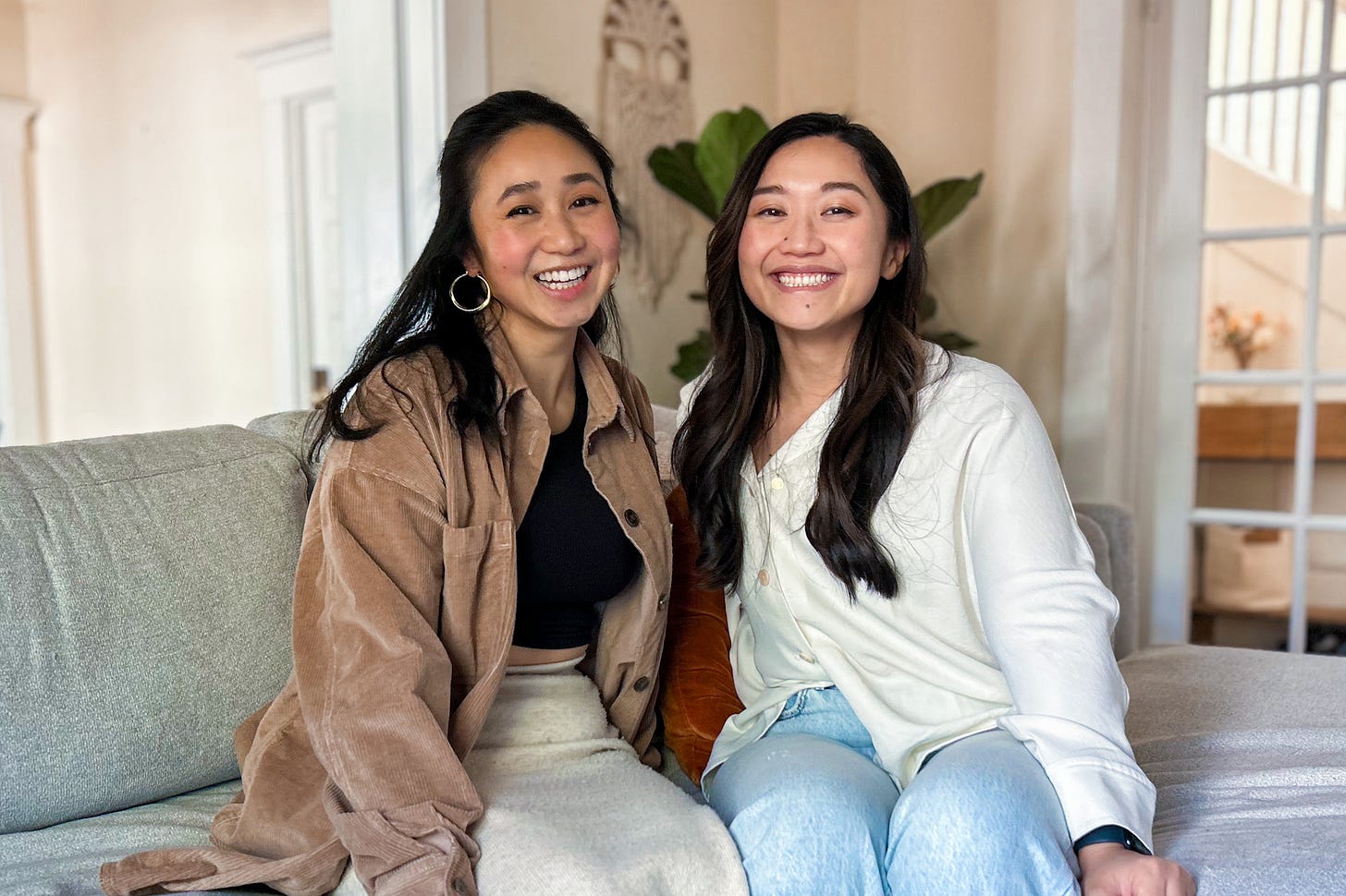 Two woman both with long, dark hair sitting on a couch and laughing towards the camera