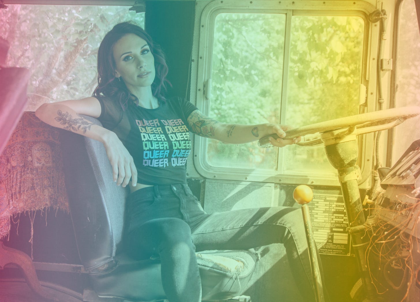 A rainow-filtered photograph of Shohreh in the driver's seat on a bus holding onto the wall with one hand with her other arm draped over the seat while wearing a shirt that says "Queer" on it repeatedly in rainbow colors