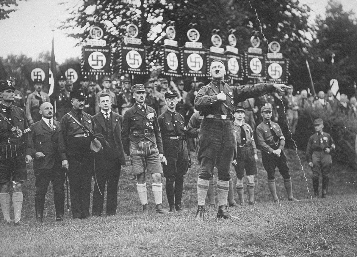 Hitler stands with his arm extended in front of a group of uniformed men. Behind them, another row of men hold banners with swastikas on them.