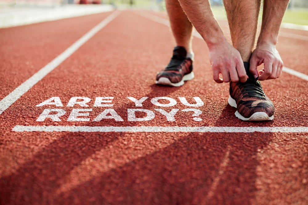 Picture of a lane of a red running track, taken at ground level looking into the distance. Text on the track says "Are you ready" and there is a person reaching down tying their shoelaces, visible from the knees down. Picture of a lane of a red running track, taken at ground level looking into the distance. Text on the track says "Are you ready" and there is a person reaching down tying their shoelaces, visible from the knees down.